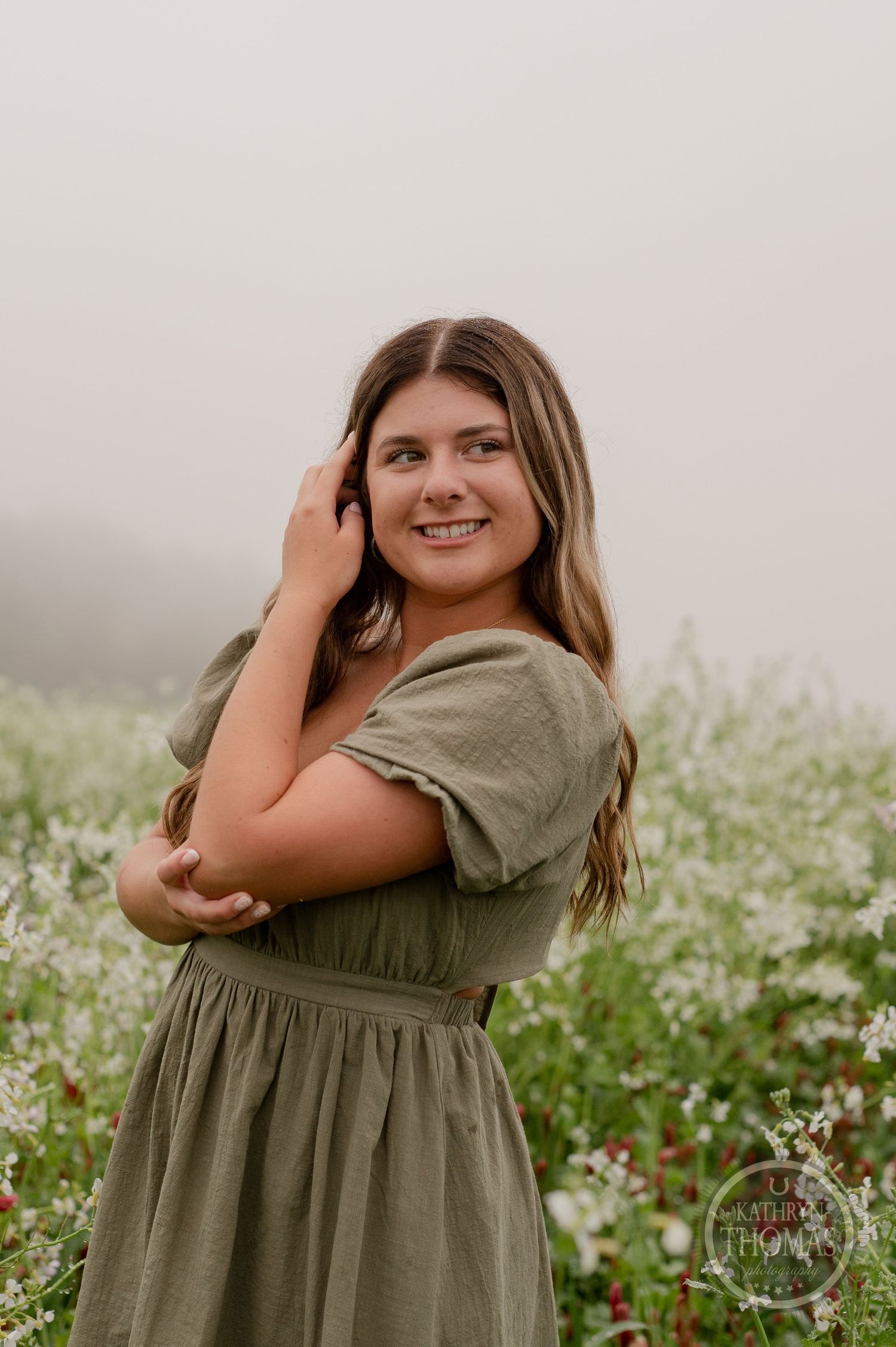 Someone in an olive-colored dress poses playfully against a backdrop of white wildflowers and misty sky.