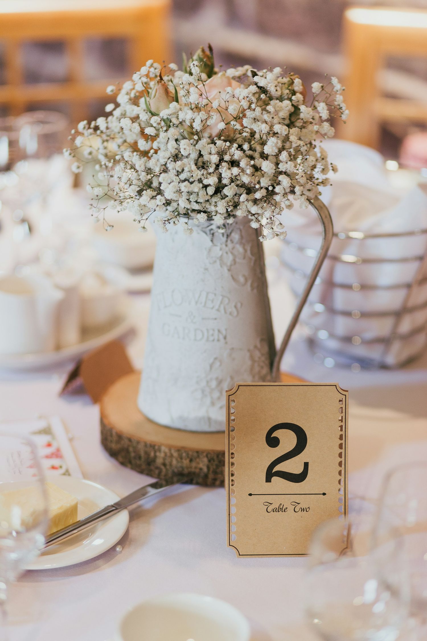Rustic wedding centerpiece featuring baby's breath flowers in a white ceramic pitcher with wooden slice base and table number.