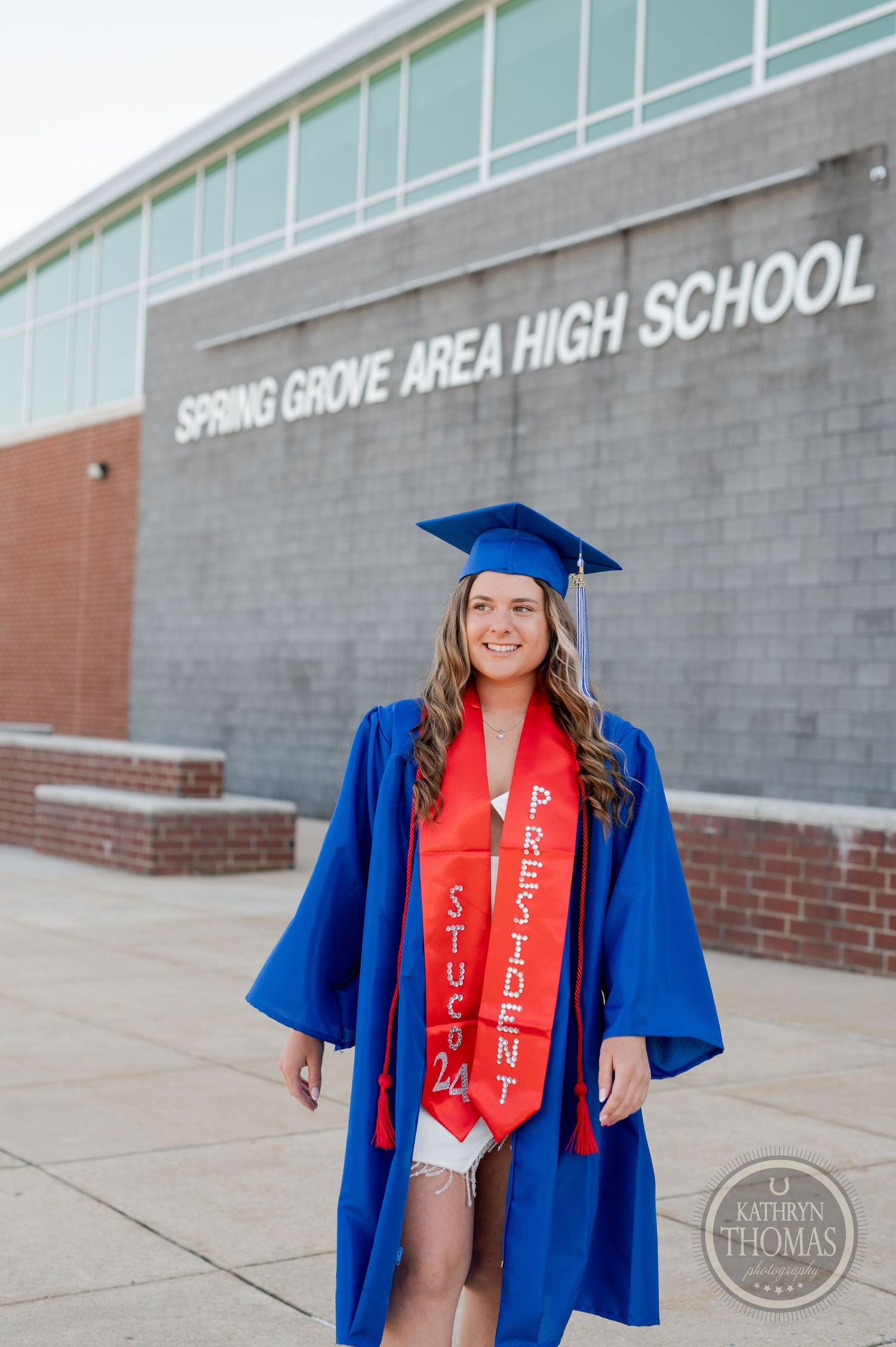 Graduate in blue cap and gown with red sash poses in front of Spring Grove Area High School building.
