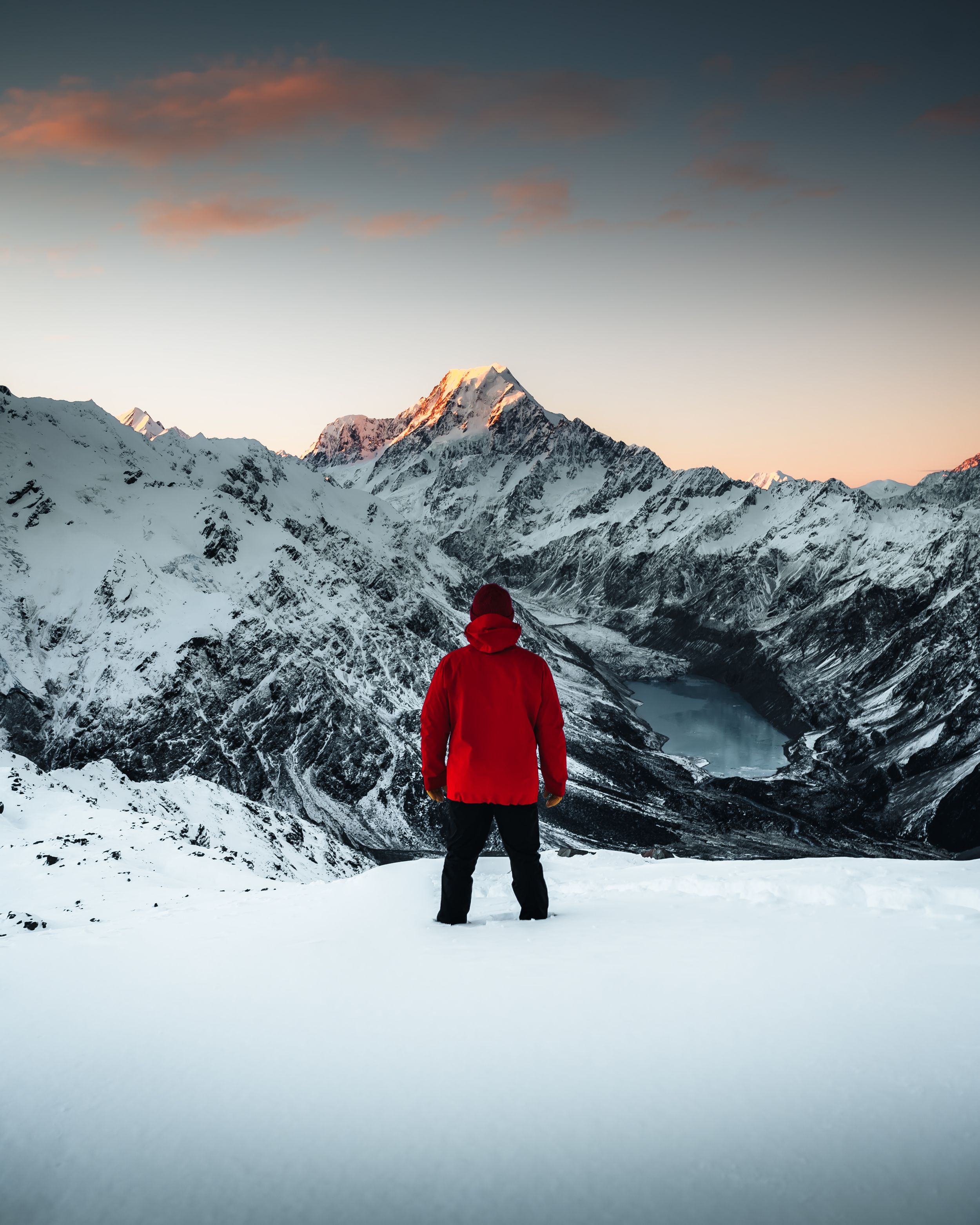 Snow-covered mountain peaks glow during sunset as a figure observes the winter landscape.