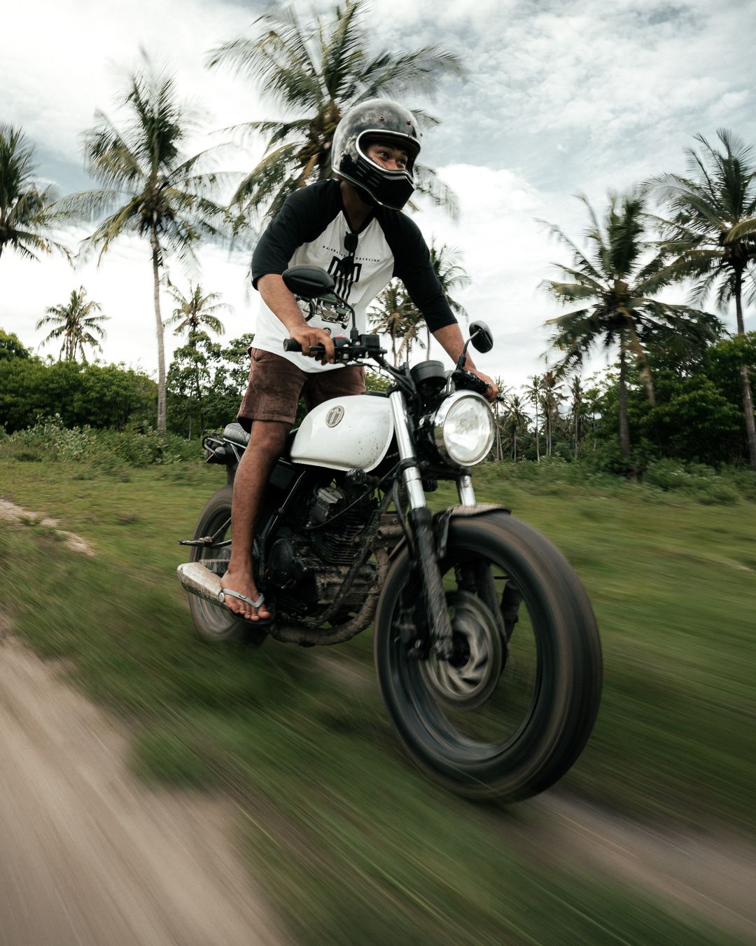 Motorcyclist riding a white vintage cafe racer bike on a tropical road with palm trees in motion blur.