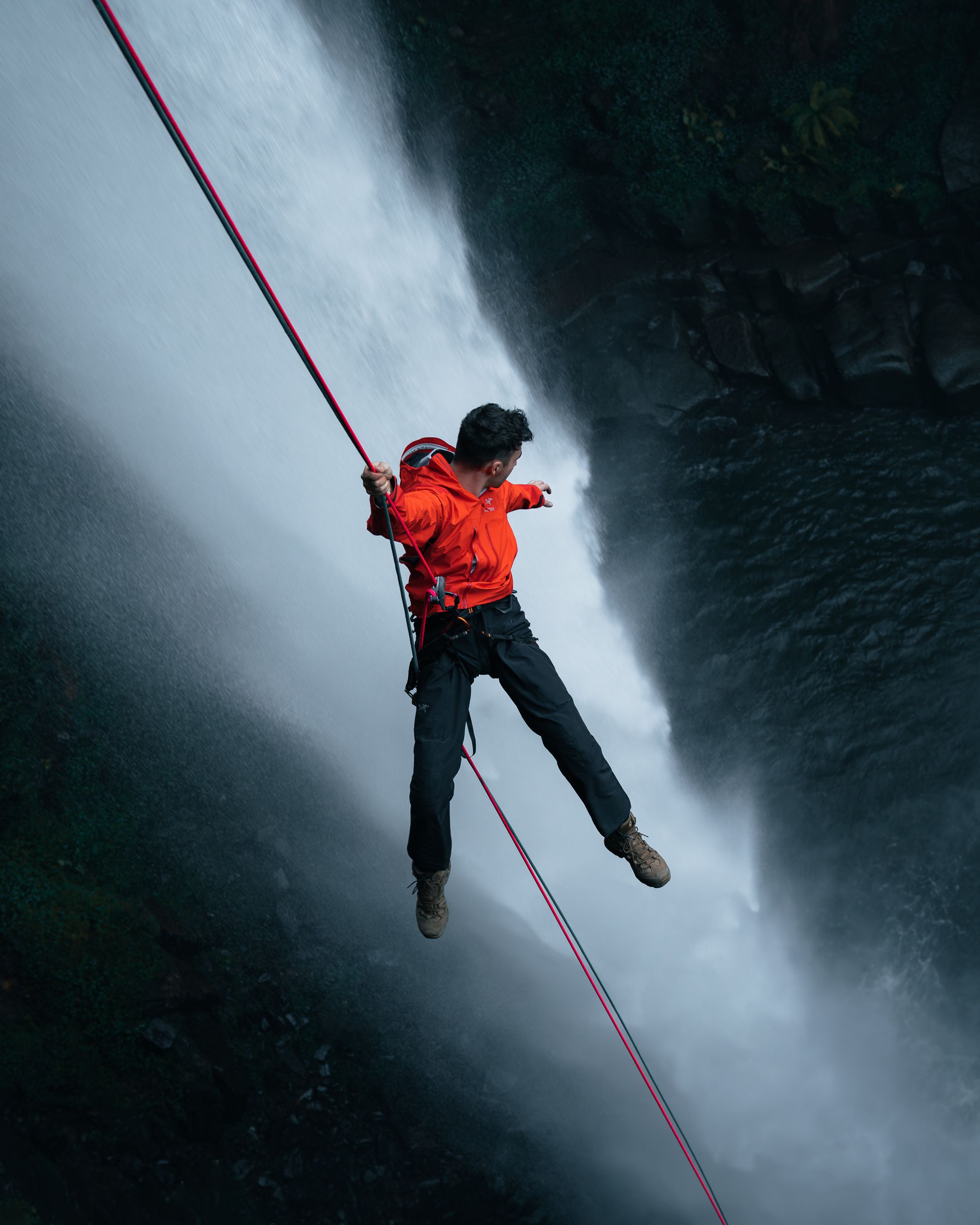 Person in red jacket rappelling down a rope against dramatic waterfall backdrop.