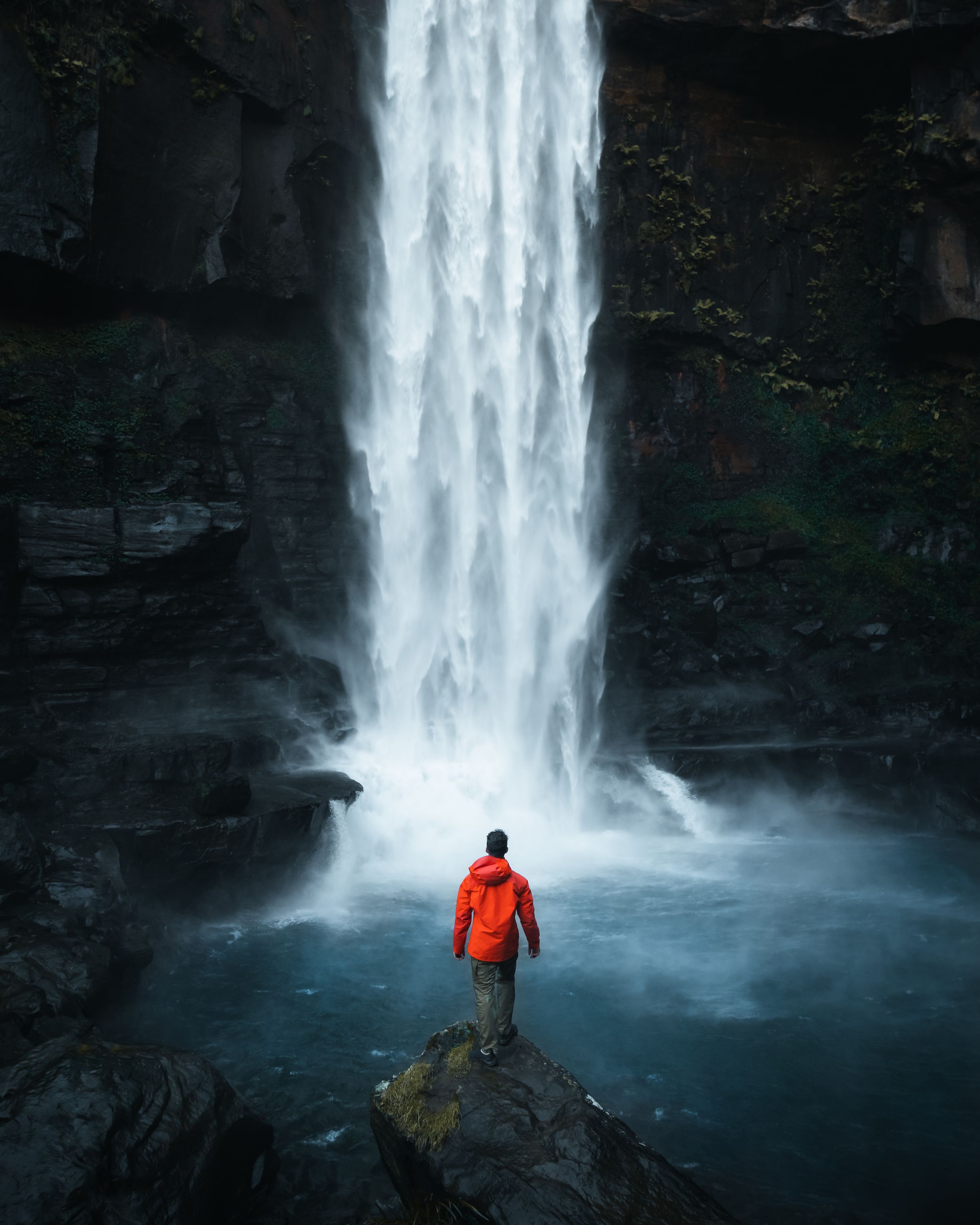 A figure in orange stands before a majestic waterfall cascading down dark rocky cliffs.