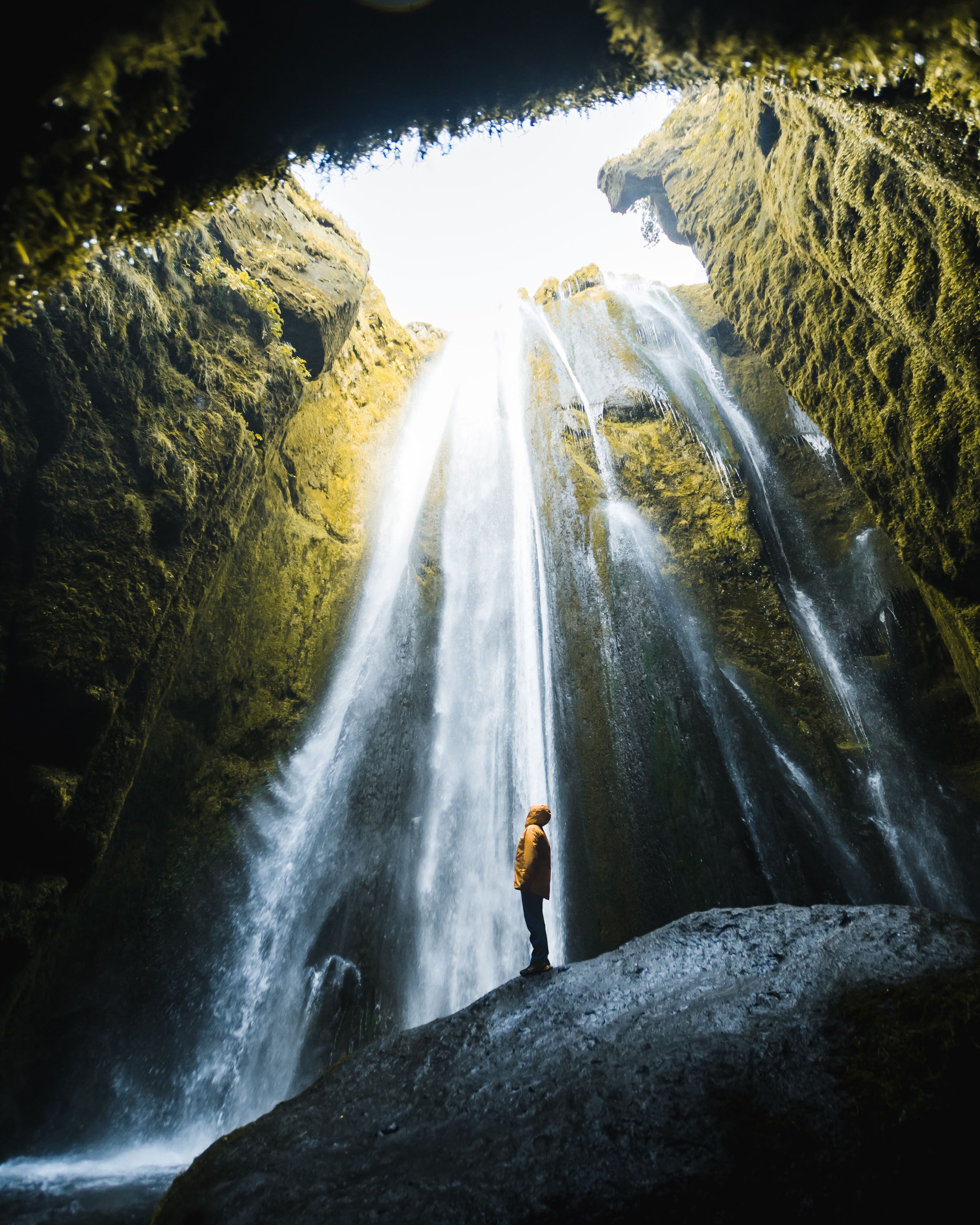 Sunlit waterfall streams down rocky cave entrance with person for scale.