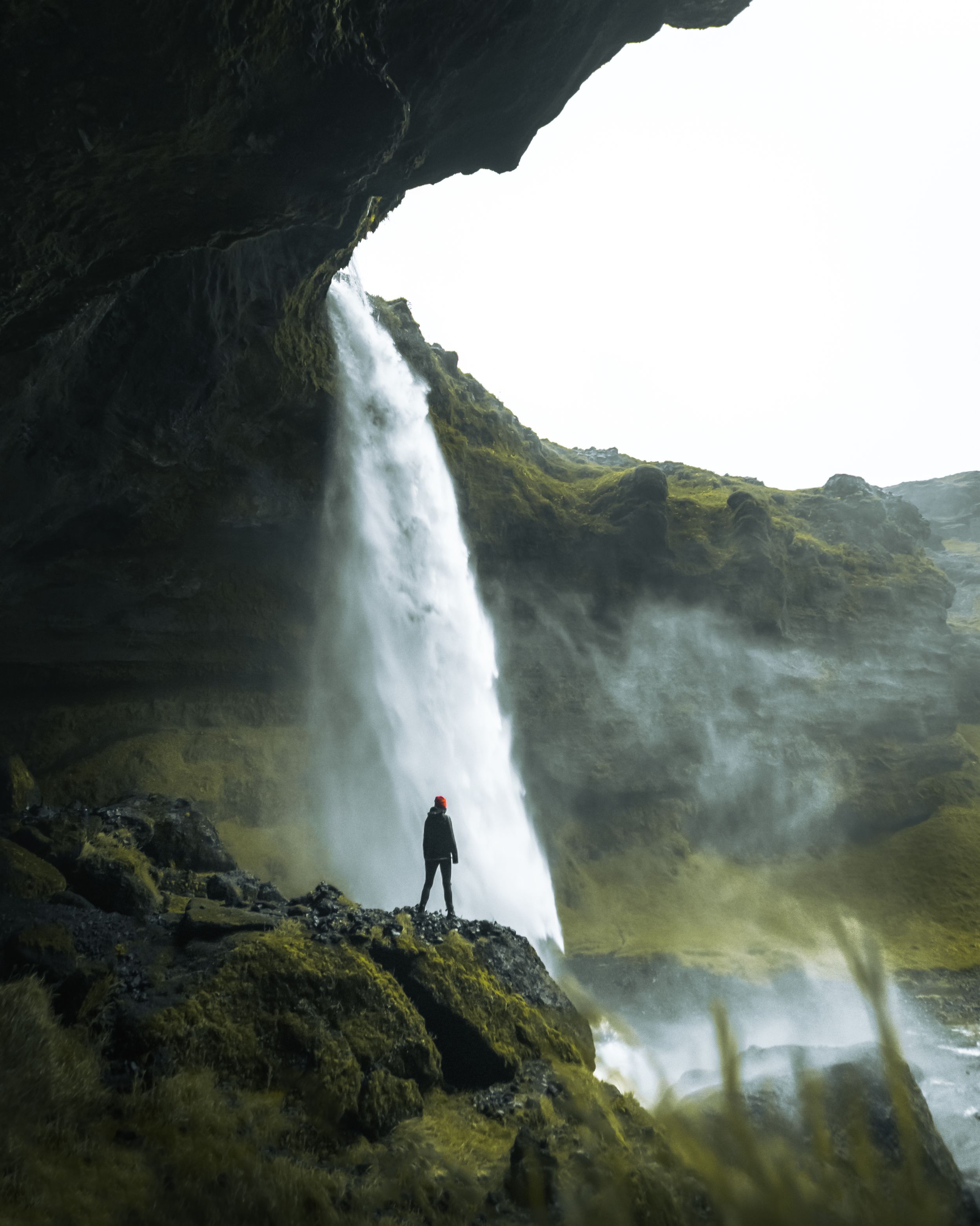 Silhouetted figure standing near powerful waterfall in mountainous terrain.