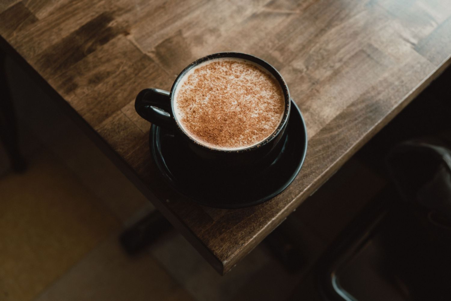 A black ceramic mug filled with creamy hot chocolate sits atop a rustic wooden table in moody lighting.