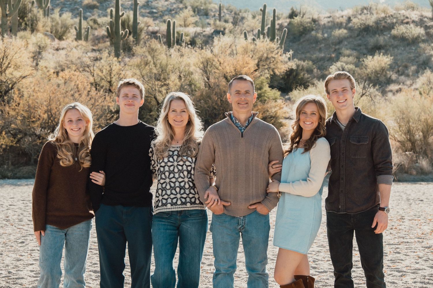 A group stands together outdoors in casual attire against a desert landscape with saguaro cacti and mountains in background.