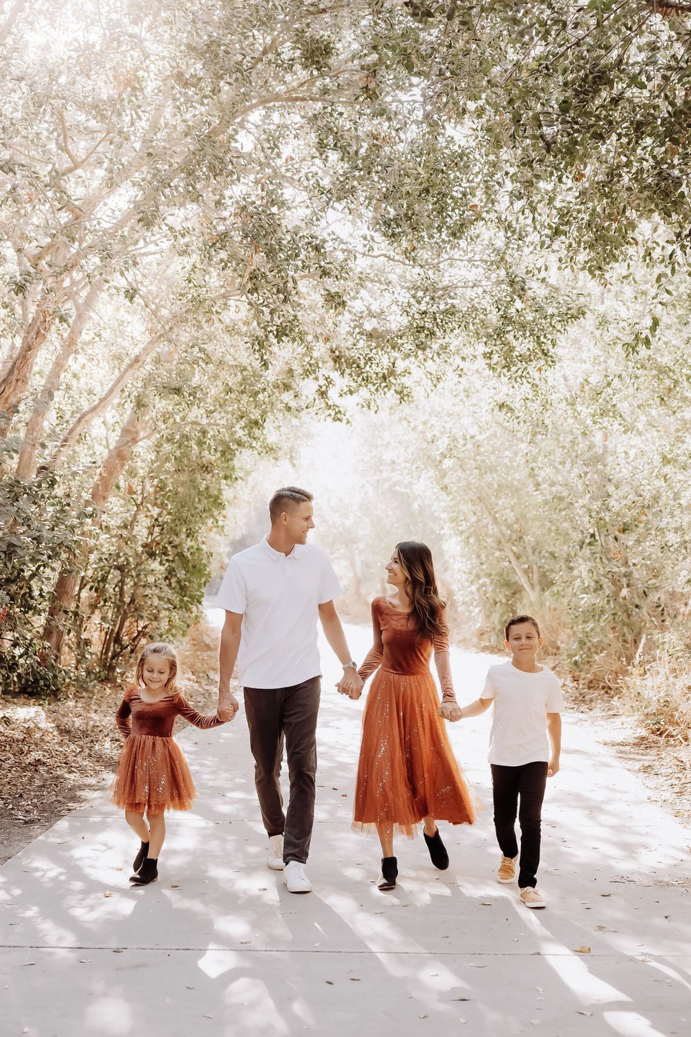 Family walking together on a sunlit tree-lined path wearing coordinated rust and white outfits in warm lighting.