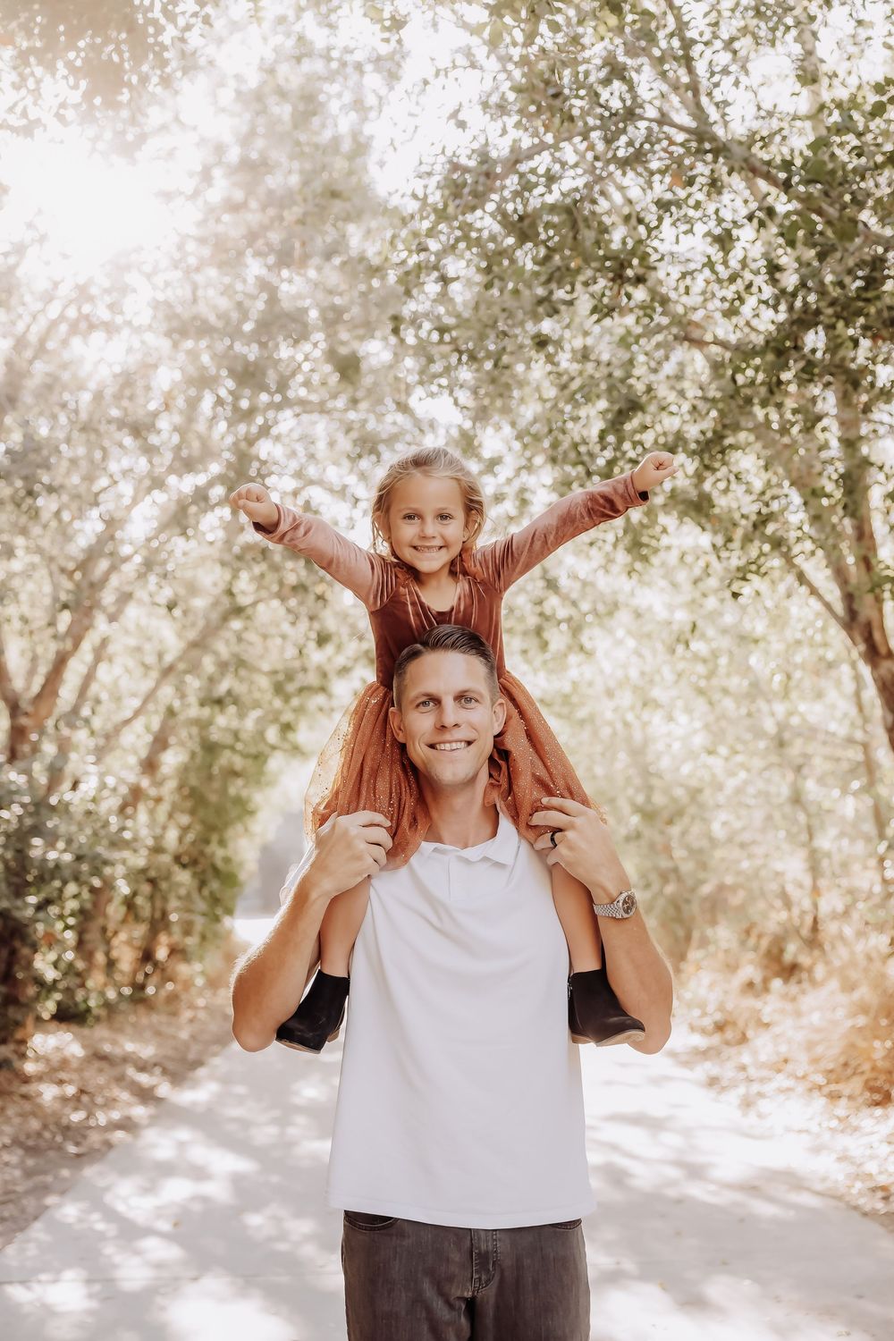 Father carries child on shoulders while playing outdoors in sunlit forest setting with trees in background.