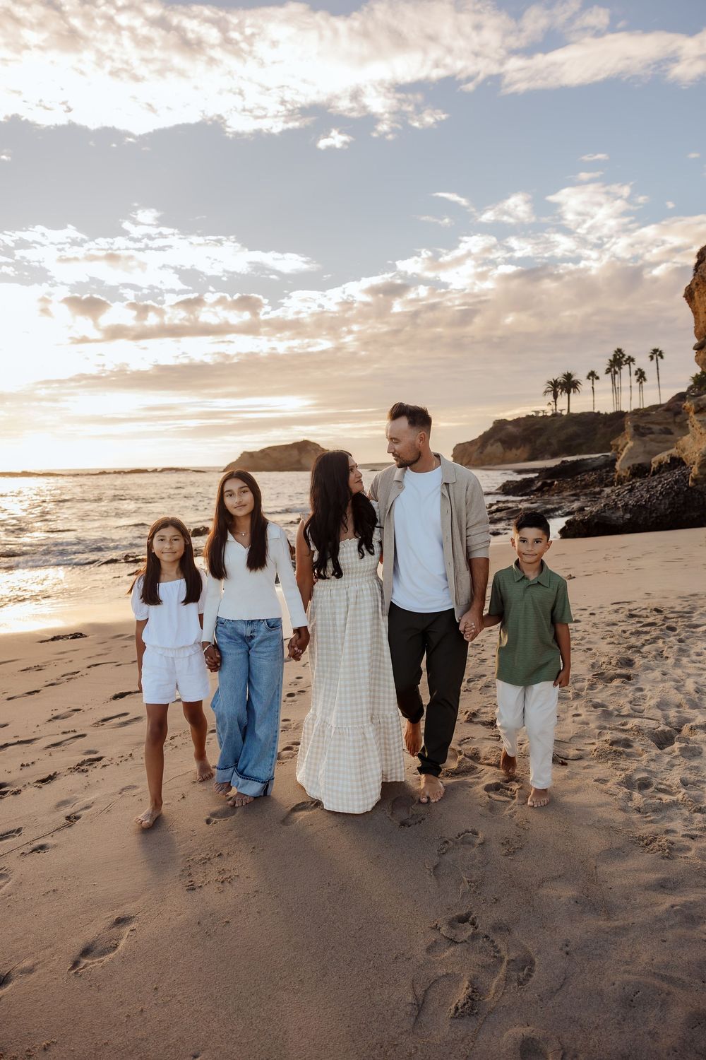 Family of five walking hand-in-hand on a sandy beach at sunset with rocky cliffs and palm trees in background.