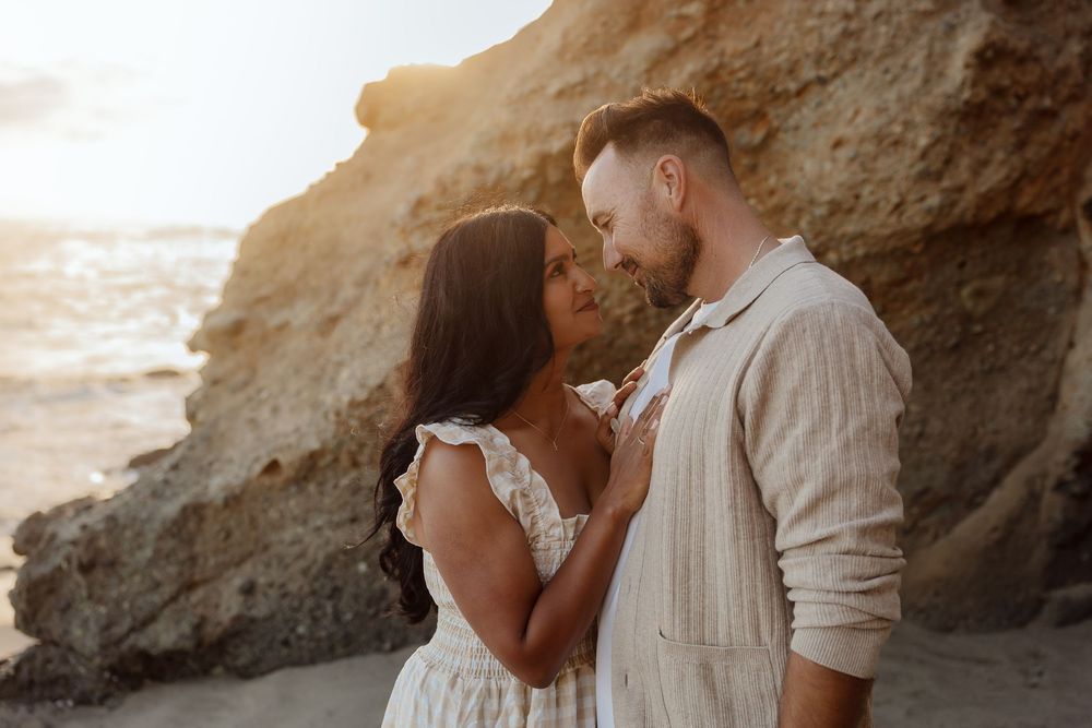 A couple shares a romantic moment on a sandy beach at golden hour, with rocky cliffs and ocean waves in the background.