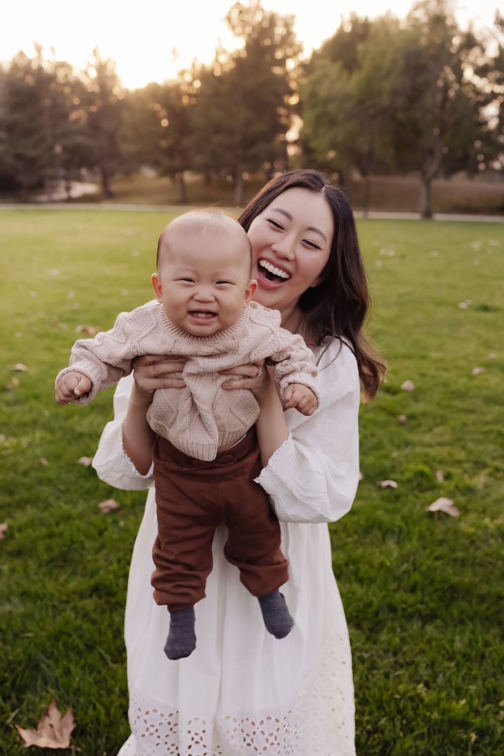 Happy mother in white dress holds laughing baby wearing brown overalls in a sunny park setting.