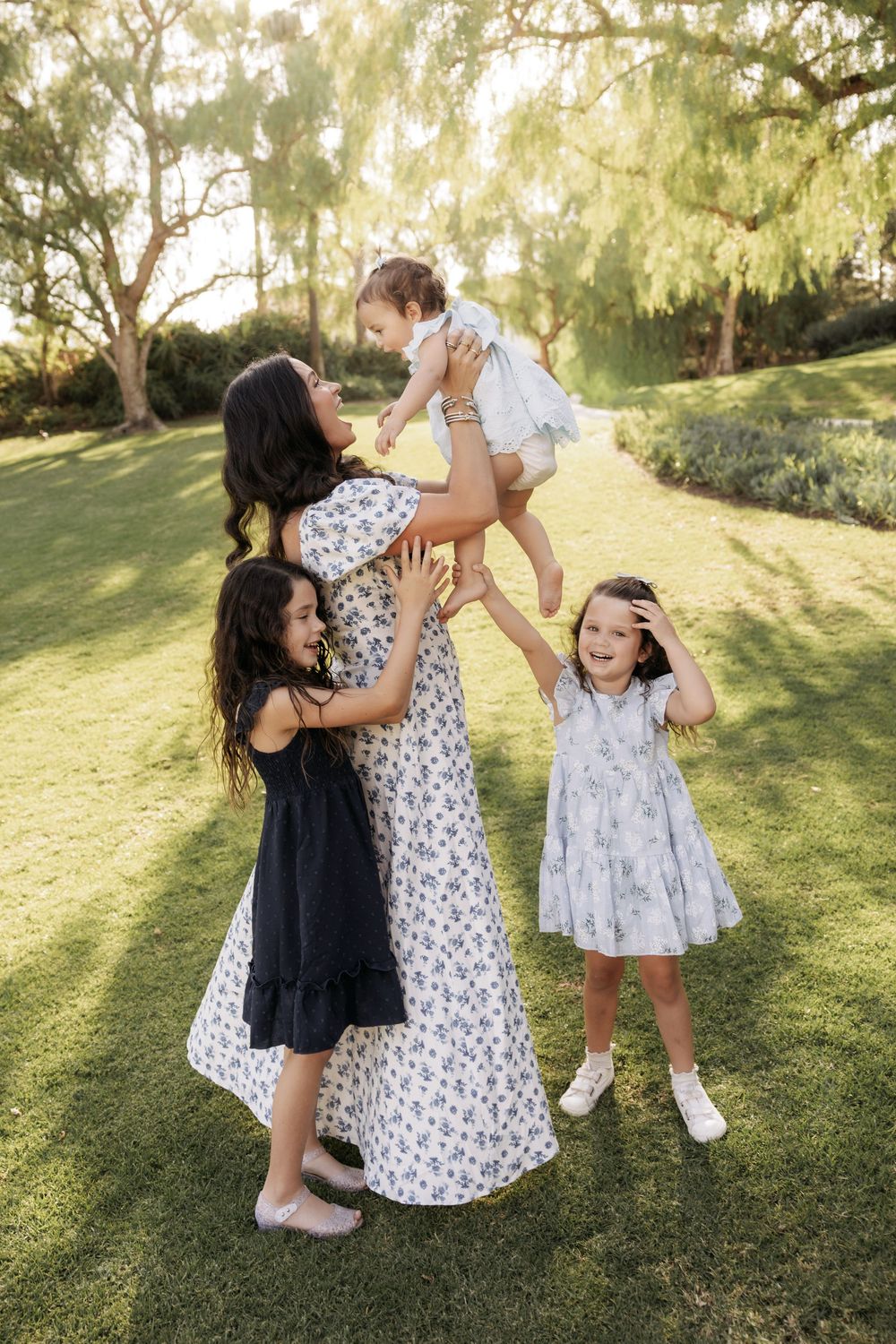 A mother lifts her baby girl while two daughters laugh joyfully in a sunlit green park.