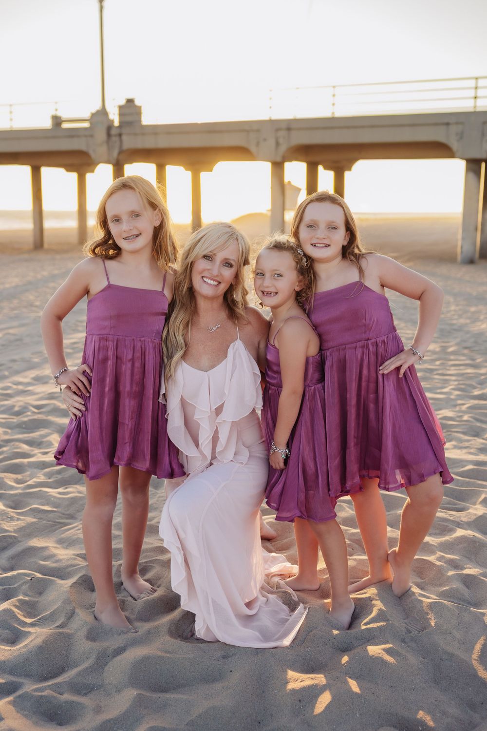 Group wearing coordinated purple and white dresses pose together on sandy beach at sunset.