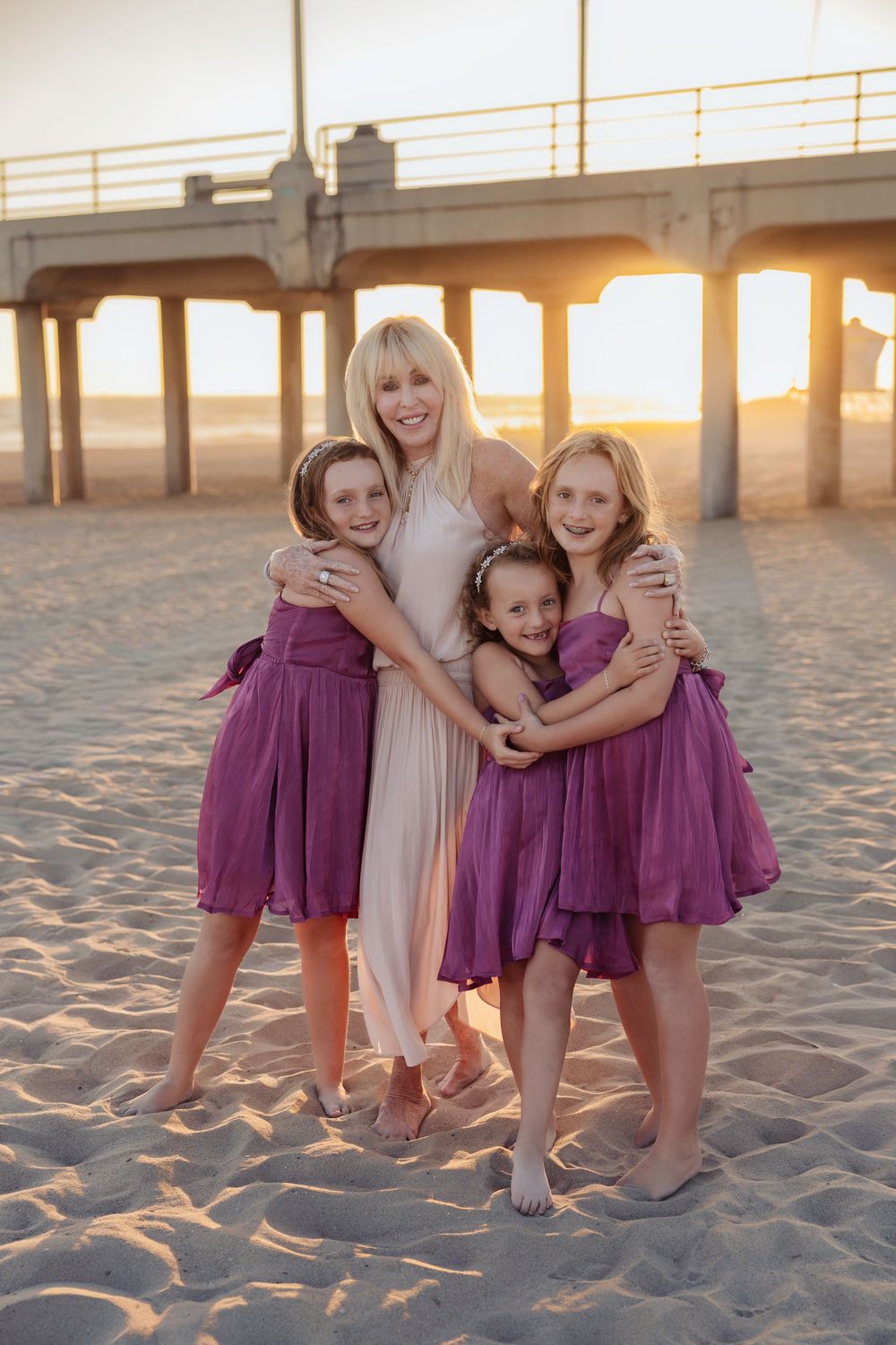Four people in coordinated dresses embrace on a sandy beach at sunset with a pier in the background.