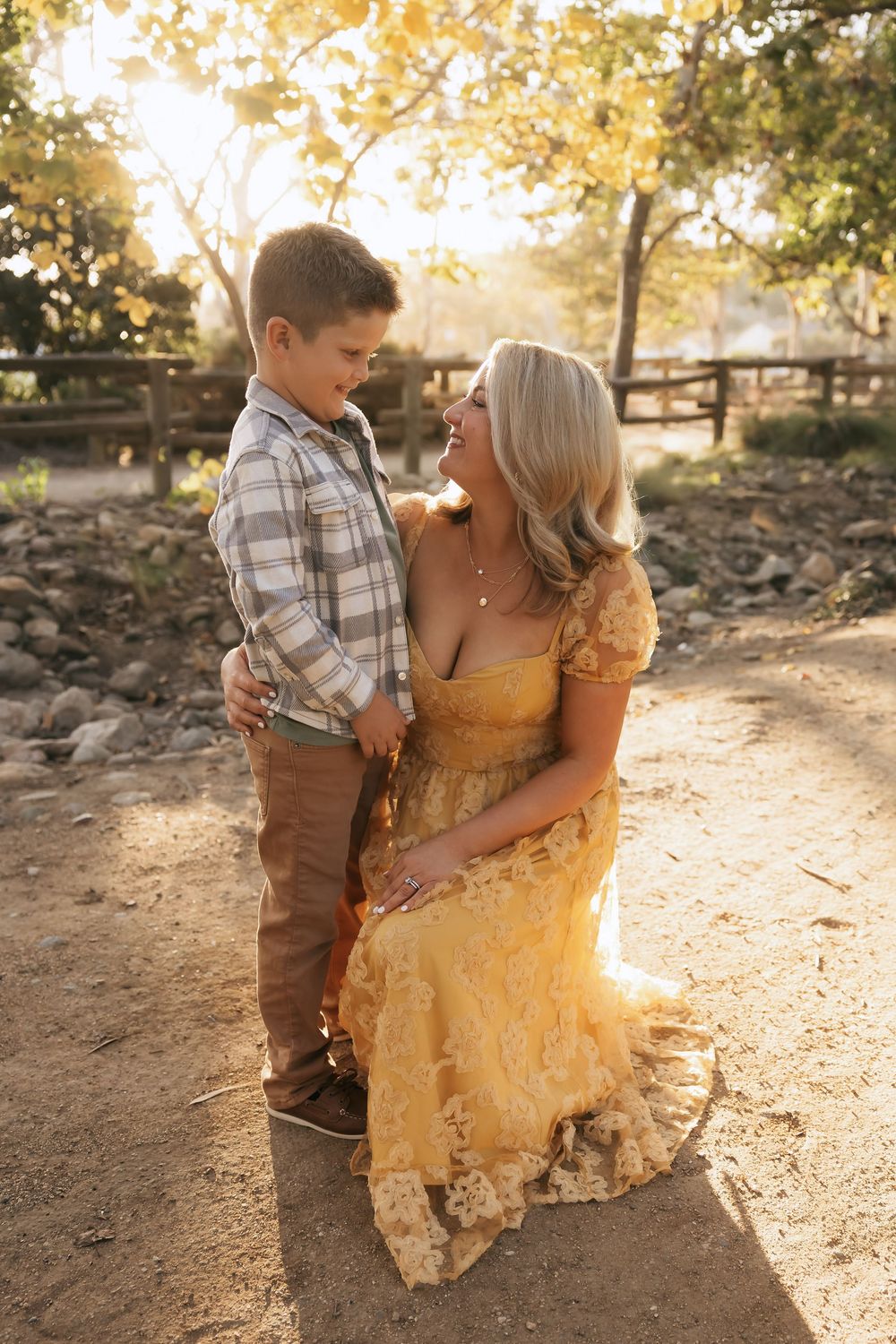 A woman in a yellow lace dress kneels beside a young boy in a plaid shirt, sharing a tender moment outdoors.
