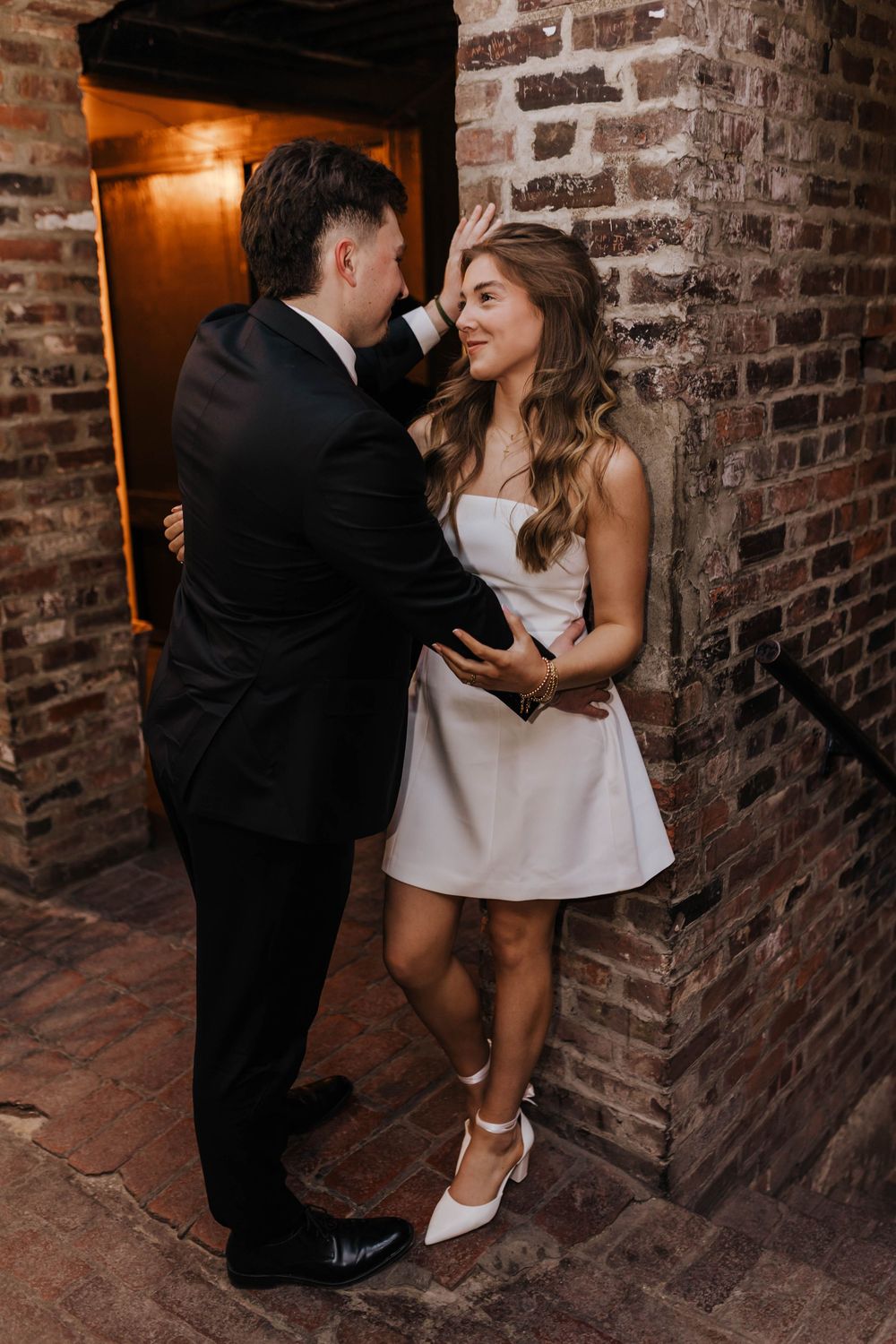 Romantic couple in formal attire share intimate moments against rustic brick walls in warm lighting.