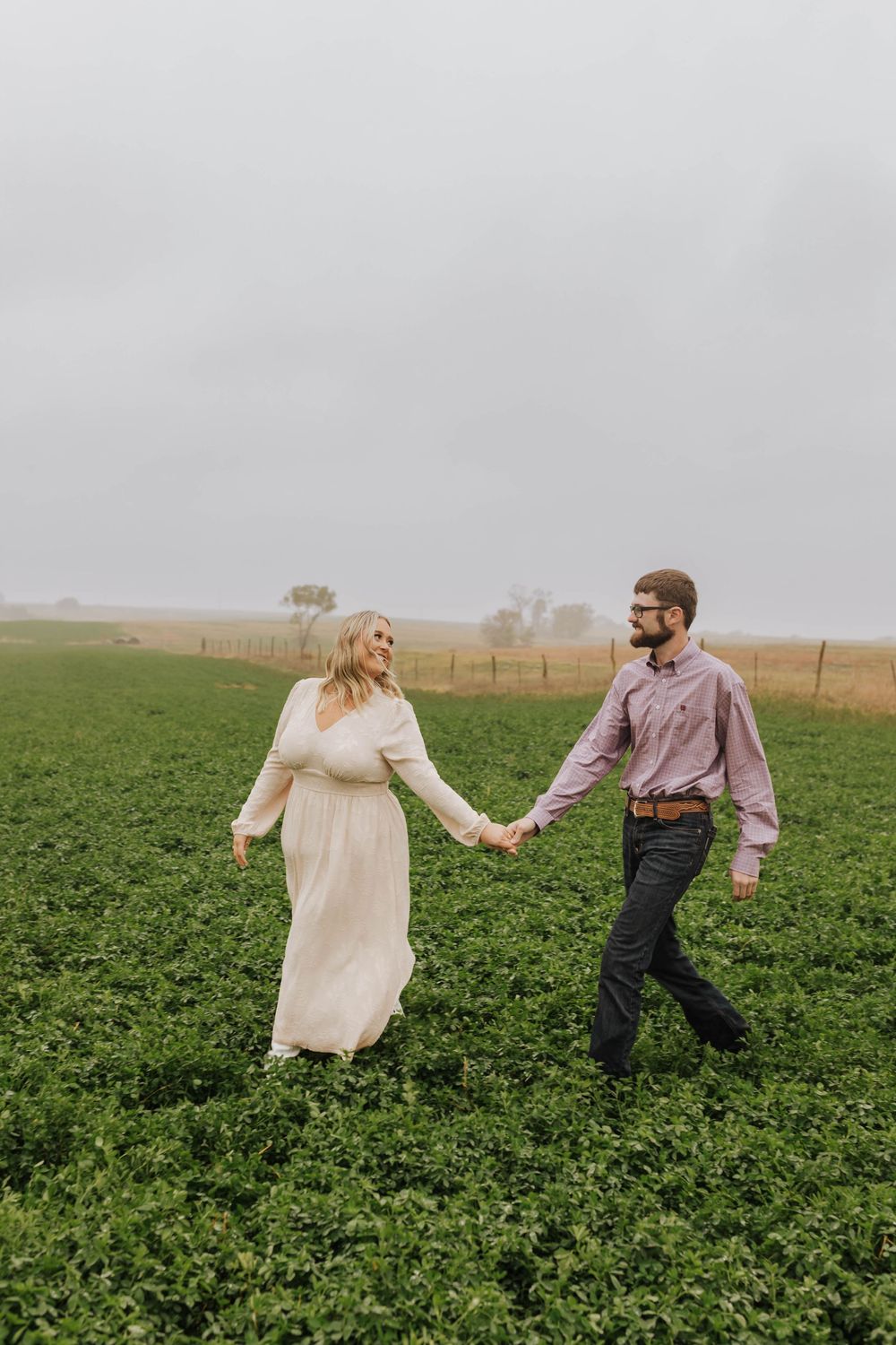 A couple runs playfully through a lush green agricultural field on a misty day.