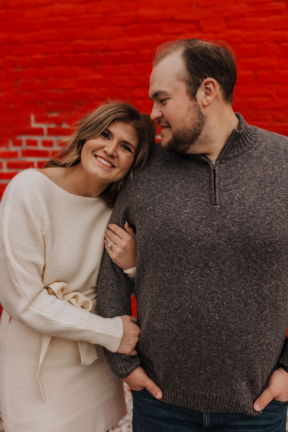 Couple in cozy sweaters share intimate moment against vibrant red brick wall.