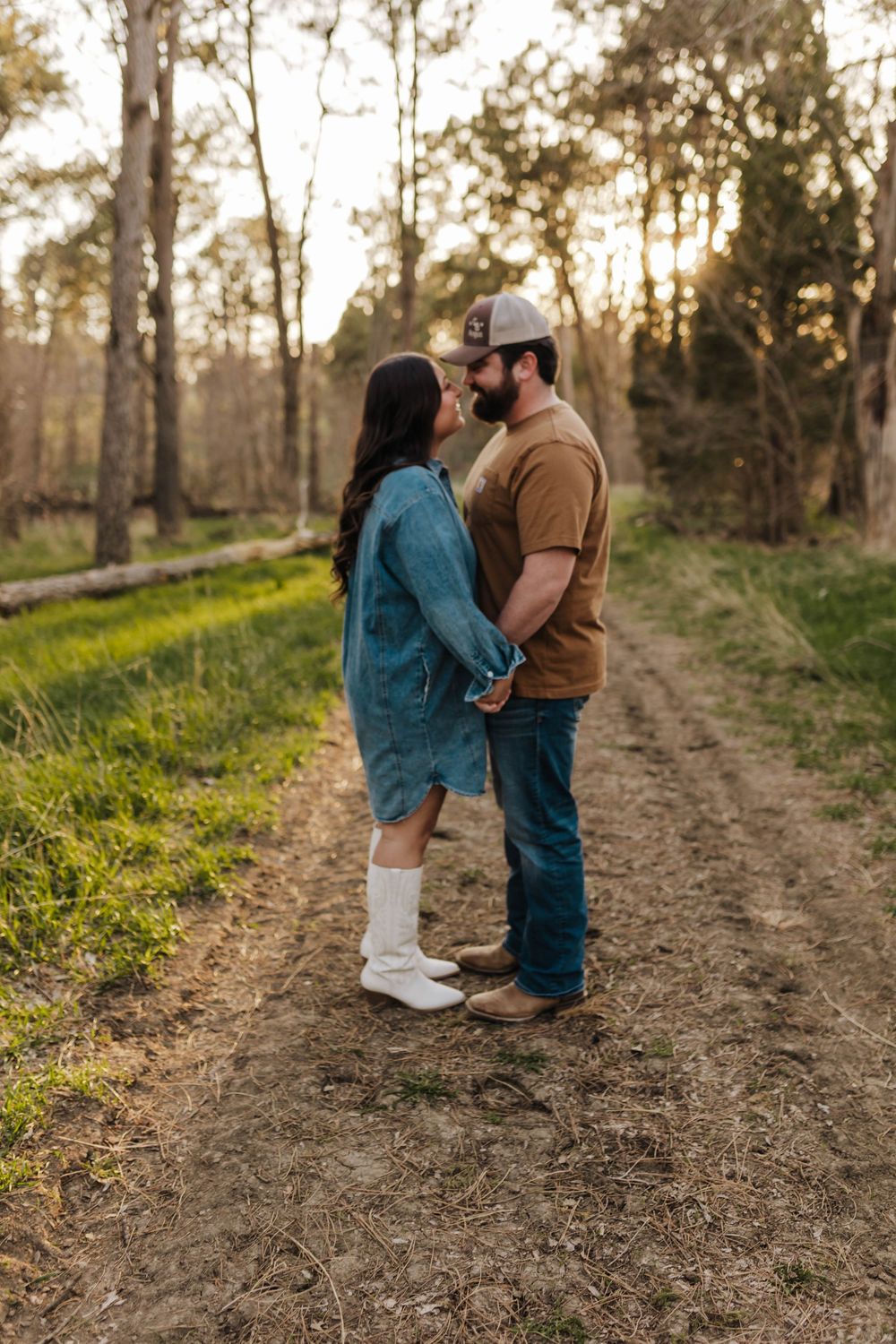 A couple shares a romantic moment on a dirt path surrounded by trees during golden hour sunset.