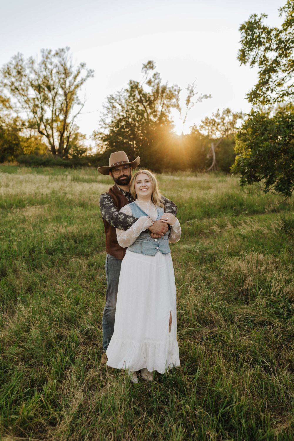 A couple embraces in a sunlit meadow at golden hour, with trees in the background.