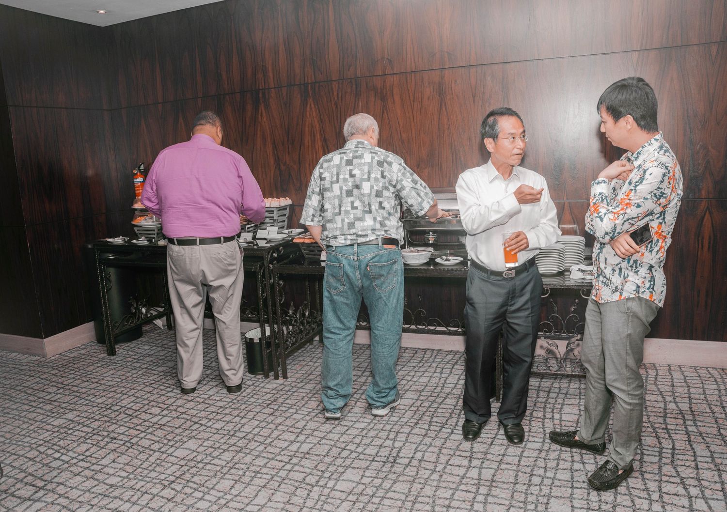 Guests in casual attire gathered around a buffet station against wood-paneled walls at an indoor event.