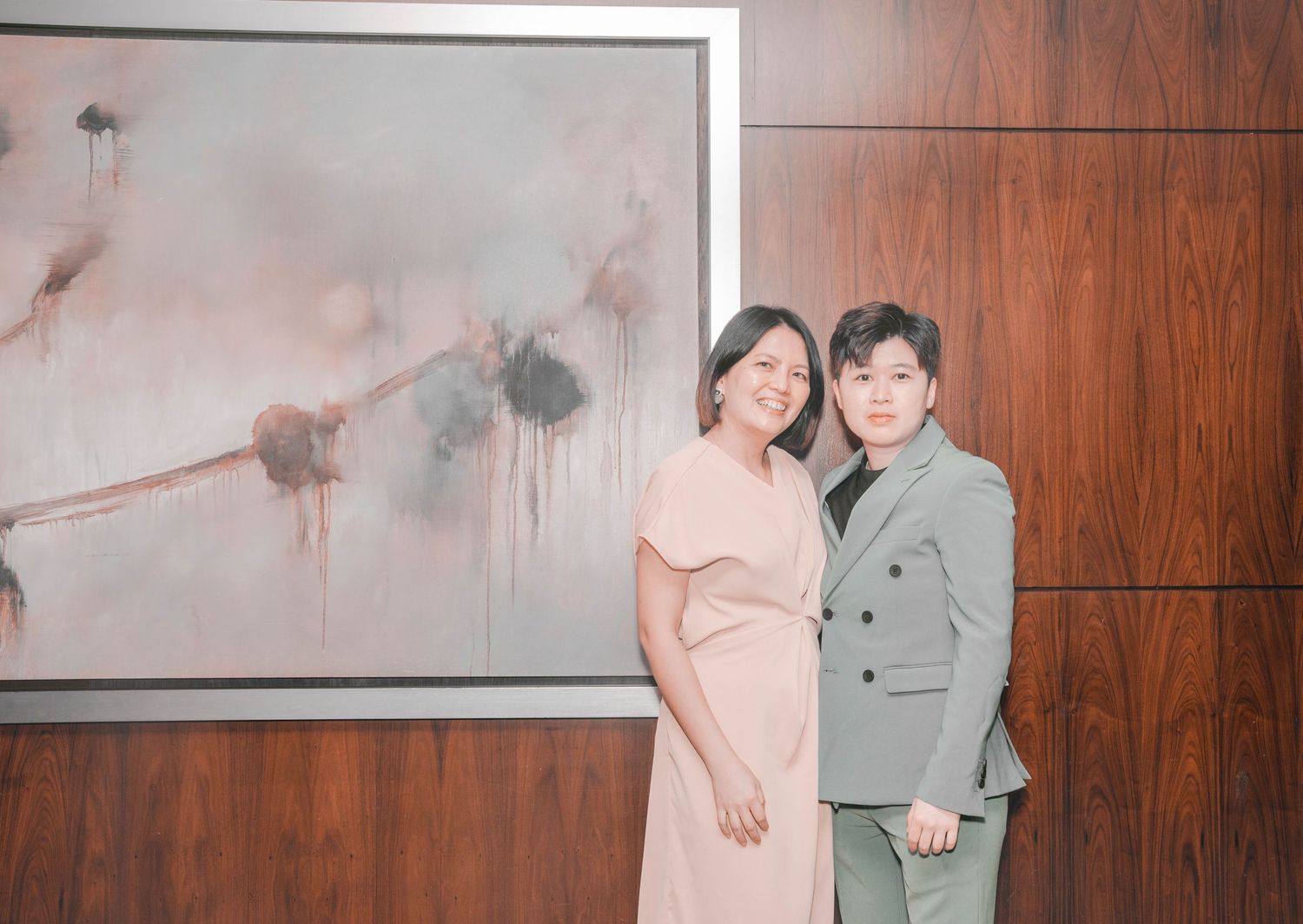 A couple poses together in front of an abstract art piece on a wood-paneled wall.