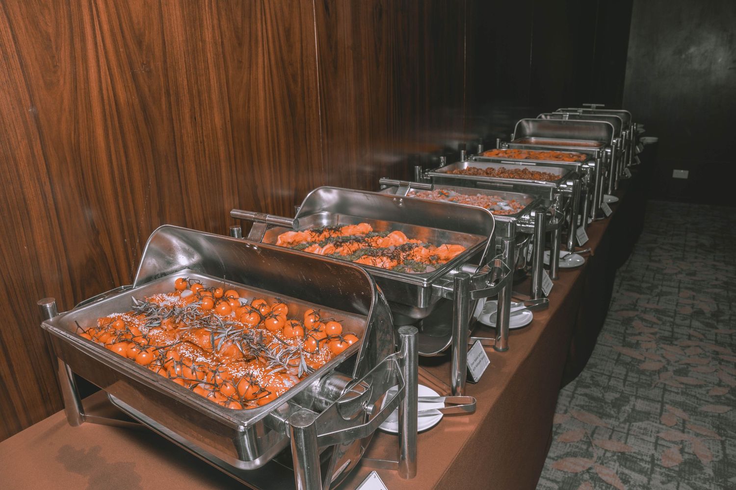 Row of stainless steel chafing dishes with orange-colored food at a buffet setup.