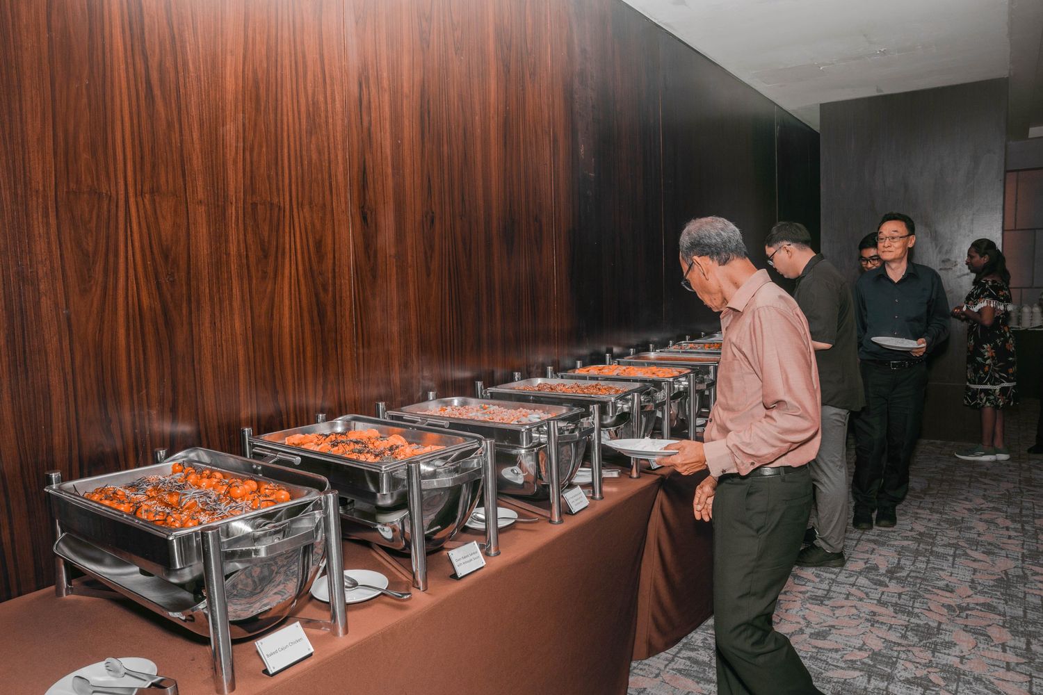 Line of buffet chafing dishes arranged against wood-paneled wall at catered event.