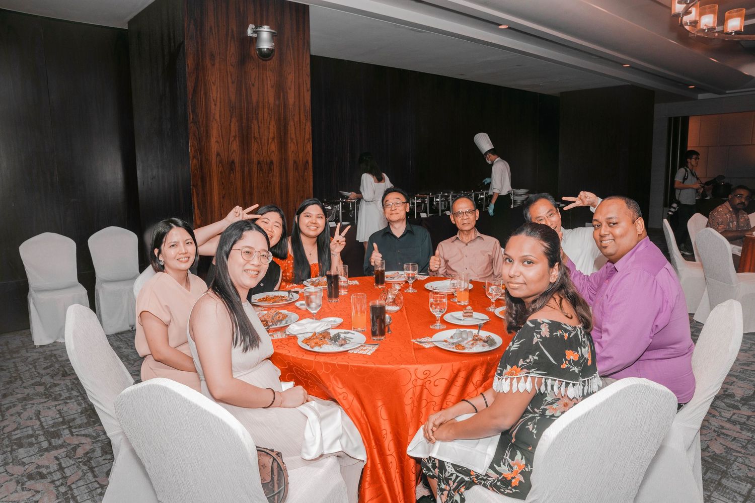Guests seated around orange tablecloth dining table at formal event venue.