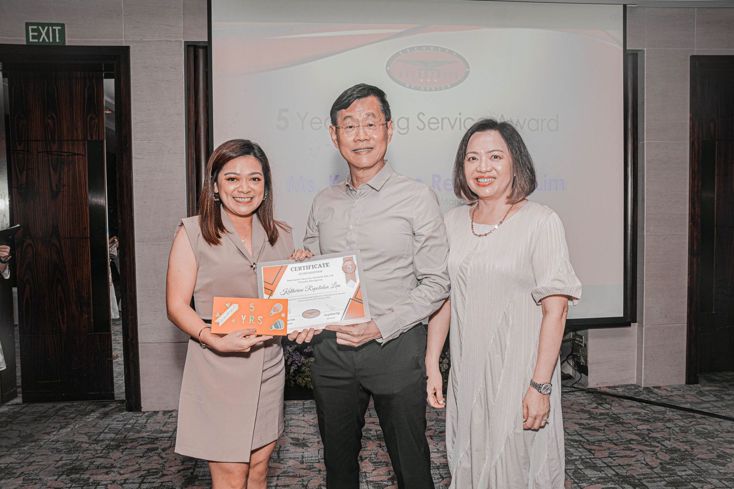 Three people pose with certificate at awards ceremony with presentation screen in background.