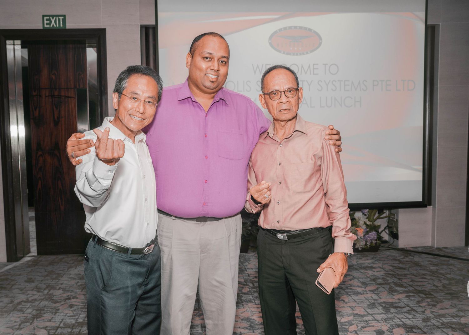 Three men posing together at a corporate event with projection screen in background.