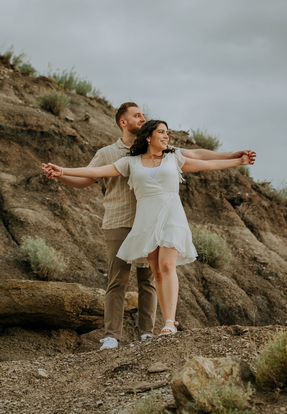 Playful couple poses with arms outstretched on a desert cliff at sunset.