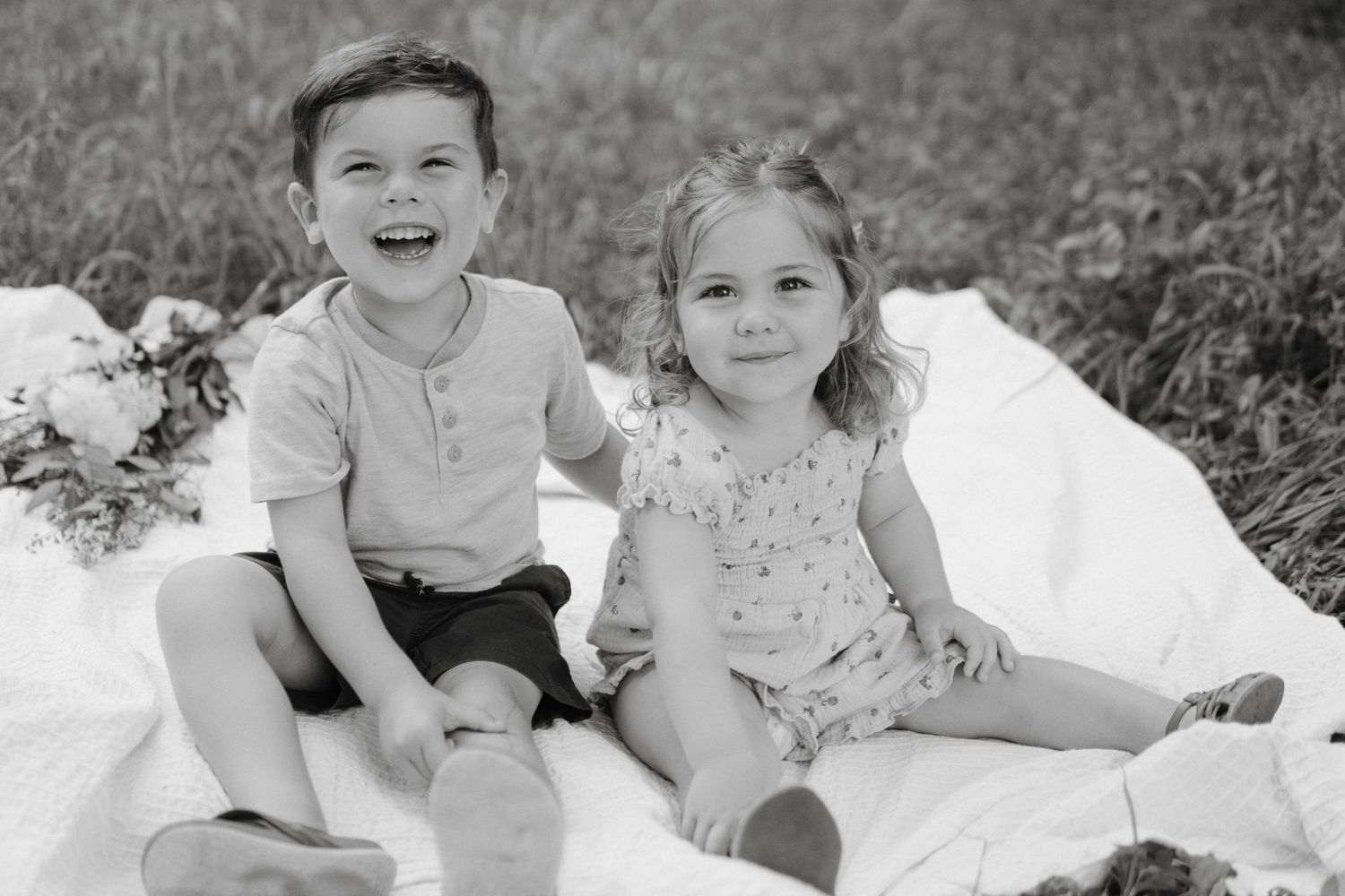 Two children sitting together on a white blanket outdoors while sharing a candid moment in black and white.