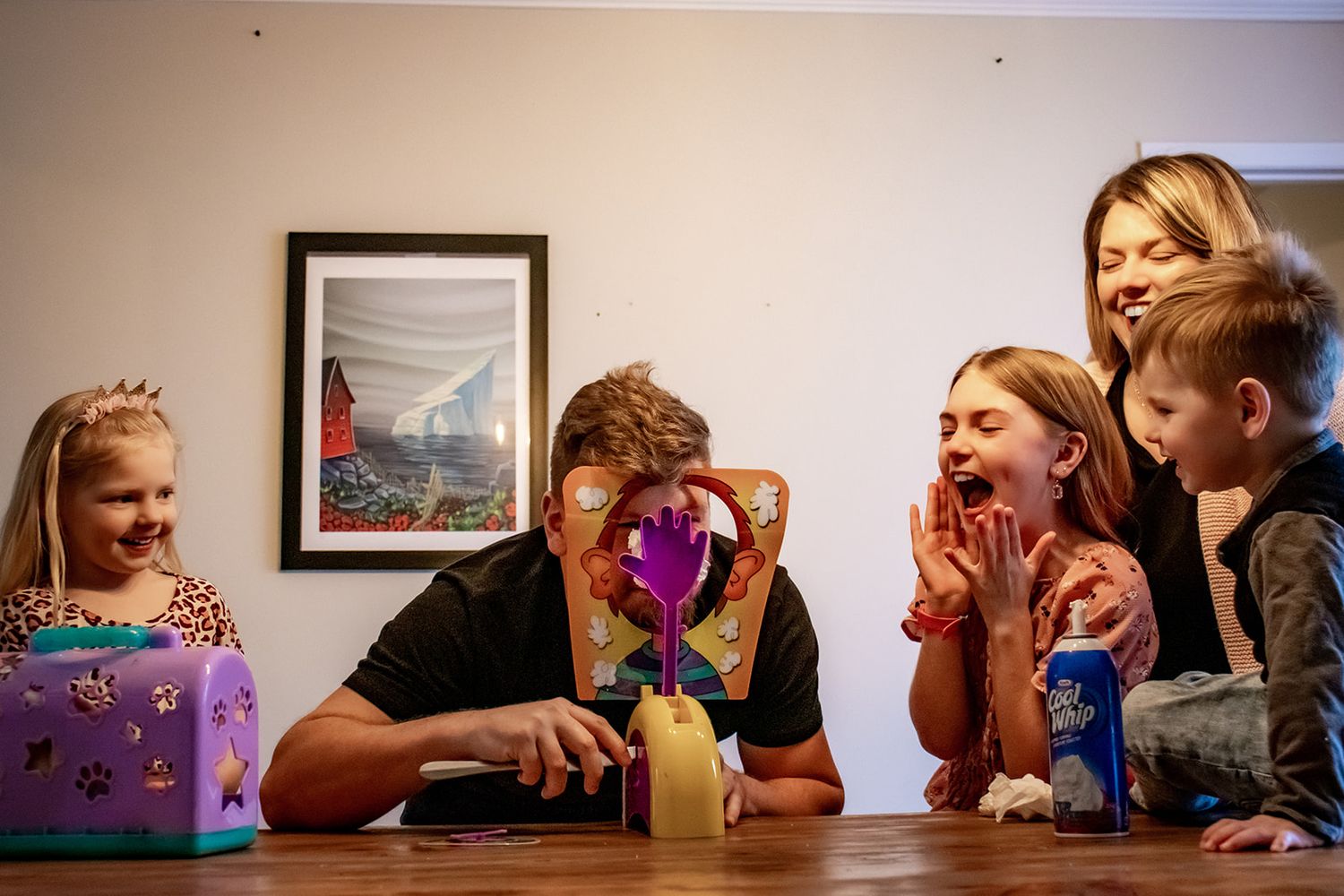 A group of people gather around a table at home, sharing laughs while looking at something on a laptop screen together.