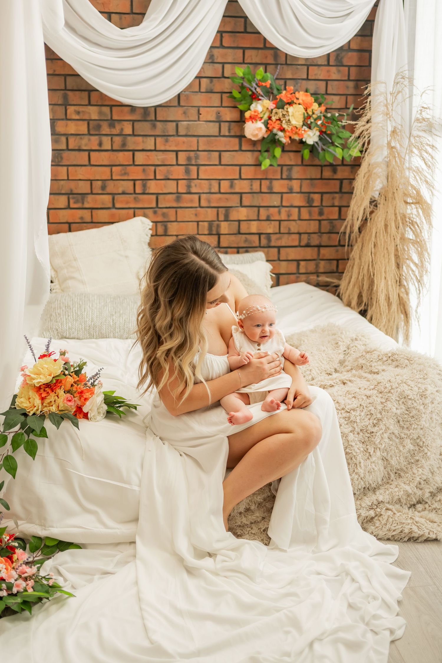 Woman in white dress sitting on bed with floral arrangements and white drapes against brick wall.