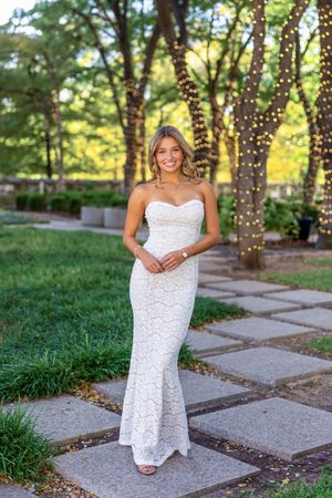 Woman in elegant white lace gown poses in garden setting with twinkling lights on trees.