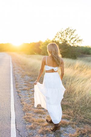 Back view of woman in flowing white dress walking along road at sunset.
