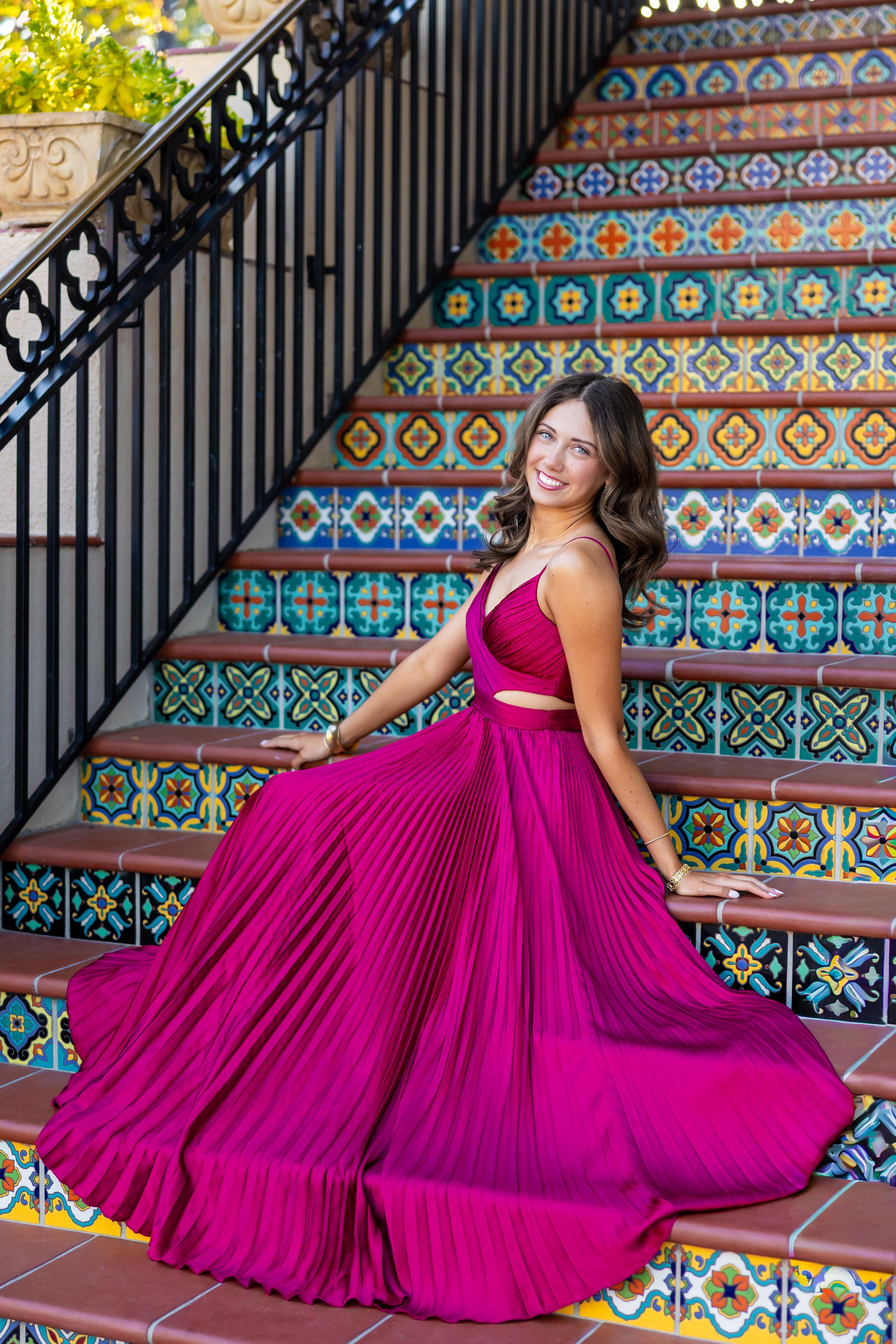 A person poses in a magenta dress on colorful tiled stairs with decorative railings.