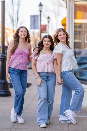 Three friends in pink tops and blue jeans pose casually together on a sidewalk outside storefronts.