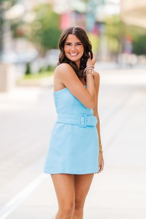 Fashion model wearing a light blue strapless mini dress poses outdoors in summer setting.