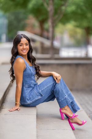 A person in denim overalls poses on concrete steps while wearing bright pink high heels.
