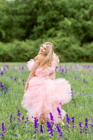 Blonde woman in a flowing pink tulle gown twirling in a field of purple wildflowers with green trees behind her.