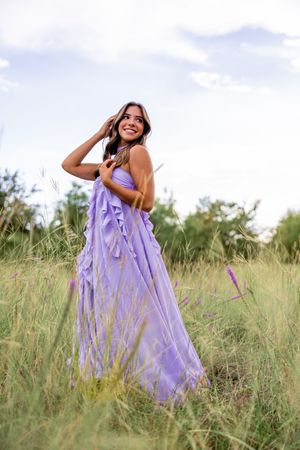 A person in a flowing lilac dress poses playfully in a sunlit meadow of tall grass during a summer photoshoot.