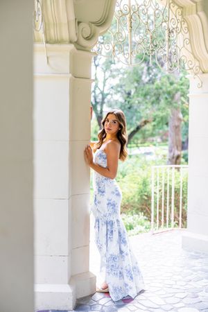 A woman in a blue and white floral sundress poses gracefully against white stone columns on a porch.