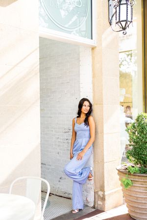 Elegant poses in blue outfit against white architectural backdrop with potted plants.