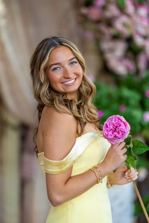 A woman in a yellow off-shoulder dress smiles while holding a pink rose against a floral background.