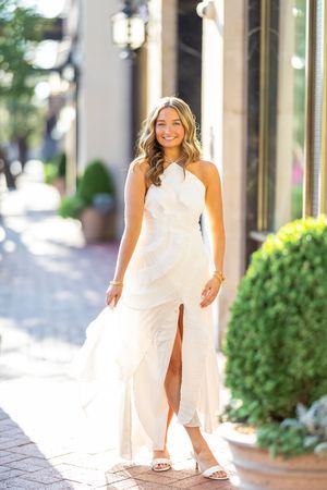 A woman in an elegant white wedding gown with a thigh slit poses on a brick sidewalk near decorative planters.