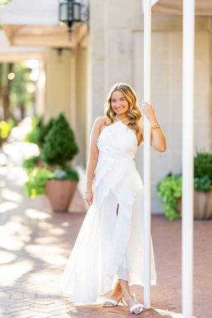 Woman in white dress poses elegantly along a covered walkway with columns and potted plants.