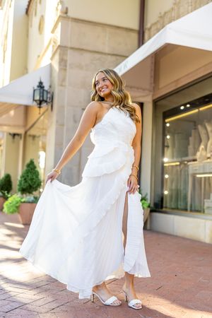A bride in a flowing white wedding dress twirls playfully on a brick patio outside an elegant building.
